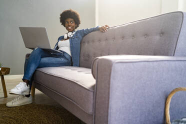 Junge Frau, die wegschaut, während sie mit ihrem Laptop auf dem Sofa zu Hause sitzt - GIOF12438