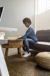 Smiling woman unboxing parcel while sitting on sofa at home - GIOF12444