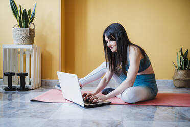 Sporty woman using laptop while sitting on exercise mat at home - EBBF03308