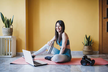 Cheerful woman holding water bottle while sitting with laptop on exercise mat at home - EBBF03310