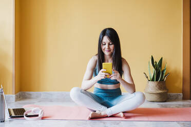 Female athlete sitting with cross legged using smart phone at home - EBBF03321