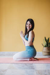 Fröhliche Frau mit verschränkten Händen übt Yoga auf einer Übungsmatte zu Hause - EBBF03326