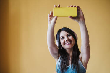 Lächelnde Frau mit Kopfhörern, die ihr Smartphone vor einer Wand zu Hause benutzt - EBBF03327