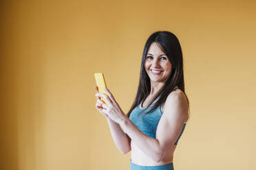 Lächelnde Frau hält Smartphone an gelber Wand zu Hause - EBBF03331
