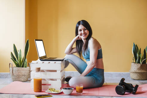 Lächelnde Frau, die zu Hause auf einer Übungsmatte am Laptop sitzt - EBBF03334