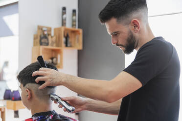 Male barber using machine to cut hair of teenage boy at salon - JRVF00411