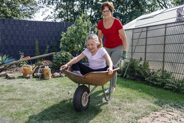 Happy girl sitting in wheelbarrow while playing with grandmother at backyard - OGF01025