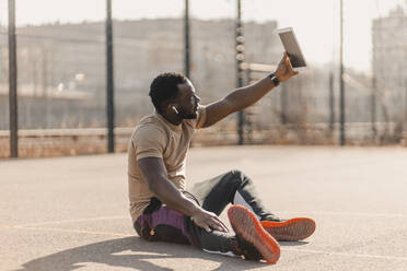 Junger Mann nimmt Selfie durch digitale Tablet beim Sitzen auf Sportplatz während sonnigen Tag - GUSF05731