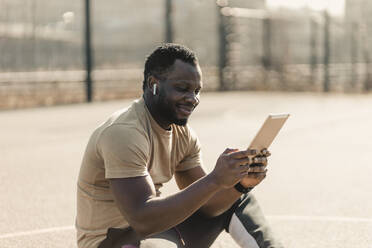 Lächelnder gut aussehender Mann, der ein digitales Tablet benutzt, während er an einem sonnigen Tag auf einem Sportplatz sitzt - GUSF05745