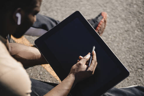 Junger Mann, der auf einem Sportplatz sitzend ein Tablet mit einem digitalisierten Stift benutzt - GUSF05749