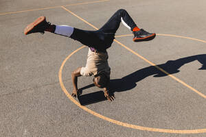 Junger Mann balanciert im Handstand mit einem Grafiktablett auf einem Basketballplatz an einem sonnigen Tag - GUSF05753