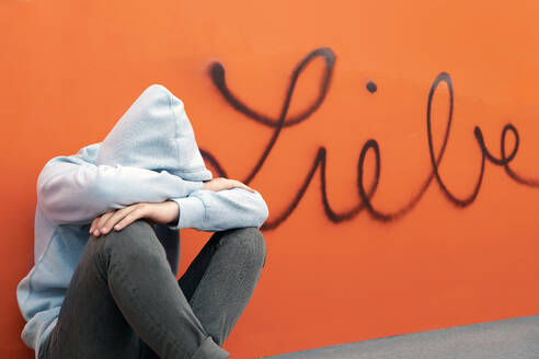 Sad teenage boy sitting in front German word for love on orange wall - PSTF00951