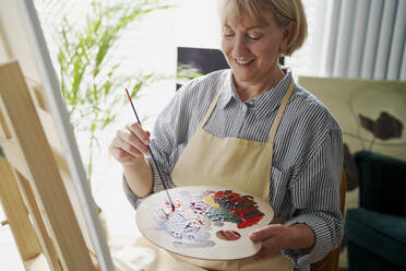 Lächelnde Frau mischt Farben beim Malen zu Hause - ABIF01360