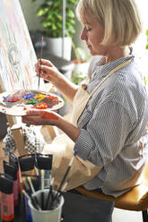 Blonde Frau mit Pinsel mischt Farben auf einer Palette vor einer Staffelei zu Hause - ABIF01362