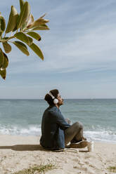 Man with eyes closed listening music through headphones while sitting at beach - AFVF08588