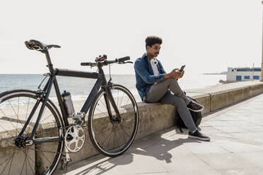 Mann, der ein Mobiltelefon benutzt, während er an einem sonnigen Tag mit dem Fahrrad auf einer Stützmauer sitzt - AFVF08594