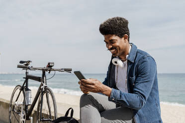 Smiling man with headphones using mobile phone while sitting on retaining wall during sunny day - AFVF08596