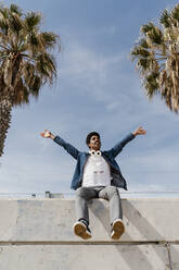 Carefree man with arms raised sitting on retaining wall - AFVF08599