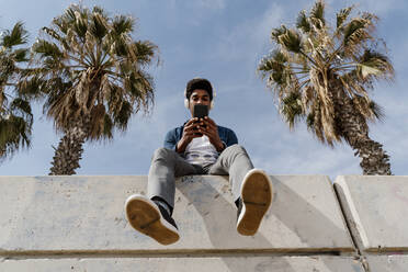 Man using smart phone while listening music through headphones on retaining wall - AFVF08601