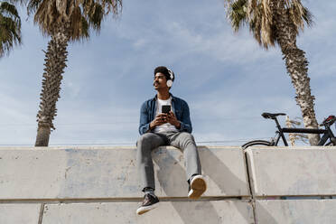 Mann in Jeanshemd schaut weg, während er sein Smartphone an einer Stützmauer hält - AFVF08602
