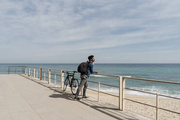 Mid adult man carrying backpack while leaning on railing by bicycle at beach during sunny day - AFVF08605