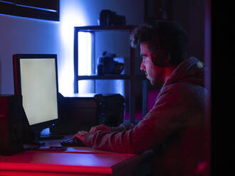Seitenansicht ruhiger junger Brünetter in Freizeitkleidung und drahtlosem Headset beim Surfen auf einem modernen Computer mit leerem Bildschirm, während er an einem Tisch in einem dunklen Raum sitzt - ADSF22547