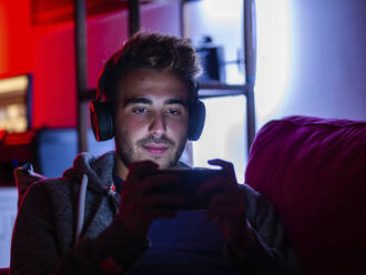 Zufriedener junger Mann in Freizeitkleidung und mit kabellosen Kopfhörern, der auf dem Sofa in einem dunklen Raum auf seinem Mobiltelefon surft - ADSF22555