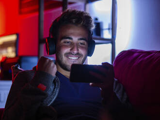 Zufriedener junger Mann in Freizeitkleidung und mit kabellosen Kopfhörern, der auf dem Sofa in einem dunklen Raum auf seinem Mobiltelefon surft - ADSF22556