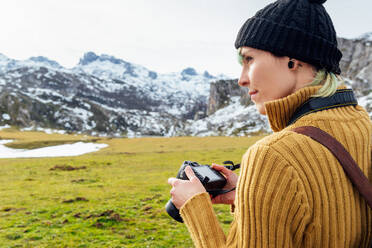 Seitenansicht fokussiert junge weibliche Fotografin in warmen Pullover nehmen Bilder auf professionelle Fotokamera von majestätischen rauen Berge auf grasbewachsenen Hochland auf klaren Herbsttag - ADSF22574