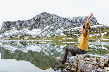 Frau mit warmem Pullover und schwarzem Hut, die mit ausgestreckten Armen auf einem rauen Stein an einem ruhigen Seeufer mit geschlossenen Augen in der Nähe einer verschneiten Felslandschaft sitzt - ADSF22579