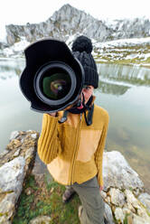 Female photographer wearing warm clothes and hat shooting pictures and looking at camera while standing on lakeside surrounded by rough snowy mountains - ADSF22580