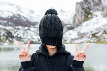 Anonyme Frau, die ihr Gesicht hinter einem schwarzen Hut versteckt und ein V-Zeichen zeigt, während sie auf einer verschneiten, rauen Hochebene am Seeufer steht - ADSF22582