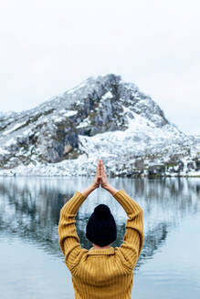 Rückenansicht unerkennbar Frau in warmer Kleidung und Hut meditiert auf Lotus Pose mit Namaste Hände am Seeufer gegen schwere verschneite Berge in Asturien - ADSF22587
