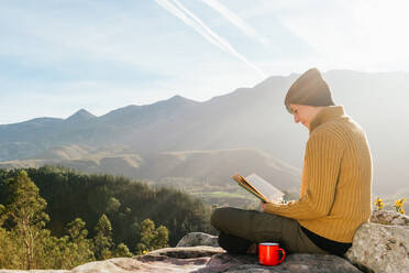 Seitenansicht eines zufriedenen weiblichen Reisenden, der mit einer Tasse Heißgetränk sitzt und ein interessantes Buch vor dem Hintergrund einer spektakulären Berglandschaft an einem sonnigen Tag liest - ADSF22590