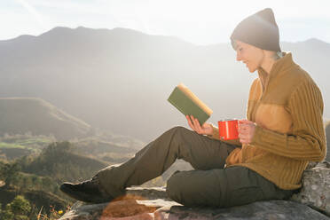 Seitenansicht eines zufriedenen weiblichen Reisenden, der mit einer Tasse Heißgetränk sitzt und ein interessantes Buch vor dem Hintergrund einer spektakulären Berglandschaft an einem sonnigen Tag liest - ADSF22592