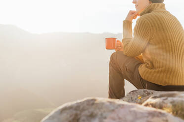 Back view of hiker sitting on stone and observing amazing scenery of highlands valley on sunny day while drinking mug of coffee - ADSF22593