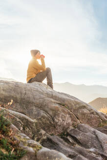 Seitenansicht eines Wanderers, der auf einem Stein sitzt und die herrliche Landschaft des Hochlandtals an einem sonnigen Tag beobachtet, während er eine Tasse Kaffee trinkt - ADSF22594