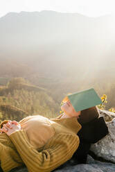 Seitenansicht eines friedlichen weiblichen Reisenden, der auf Felsen liegt und sein Gesicht mit einem Buch bedeckt, während er in den Bergen an einem sonnigen Tag schläft - ADSF22597