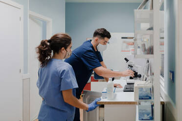 Side view of crop anonymous male medic in uniform and mask using microscope while working in lab near woman colleague - ADSF22624