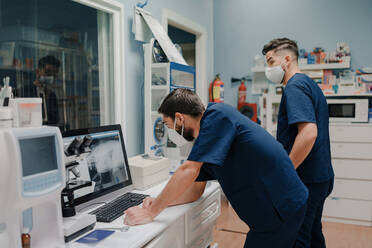 Anonyme junge männliche Ärzte in Atemschutzmasken und Uniformen bei der Arbeit an einem Desktop-Computer in einer Klinik - ADSF22625