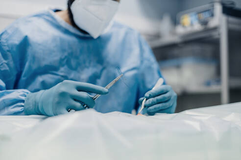 Anonymer männlicher Tierarzt in Uniform und Atemmaske, der während einer Operation im Krankenhaus medizinische Instrumente benutzt - ADSF22633