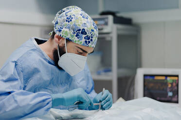 Focused male veterinarian in uniform and respiratory mask using medical instruments during surgery in hospital - ADSF22634