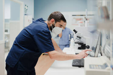 Seitenansicht eines anonymen männlichen Mediziners in Uniform und Maske, der ein Mikroskop bei der Arbeit im Labor benutzt - ADSF22637