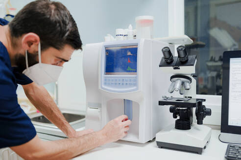 Seitenansicht eines anonymen männlichen Arztes mit Atemmaske, der eine professionelle Ausrüstung mit Diagrammen in der Nähe des Mikroskops im Labor benutzt - ADSF22638