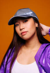 Porträt einer asiatischen jungen Frau mit ernster Miene, die eine lila Jacke und eine graue Mütze trägt und vor einem gelben Hintergrund in die Kamera blickt, während sie ihren Kopf berührt. - ADSF22692