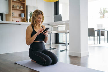 Woman using smart phone while kneeling on exercise mat at home - MPPF01639