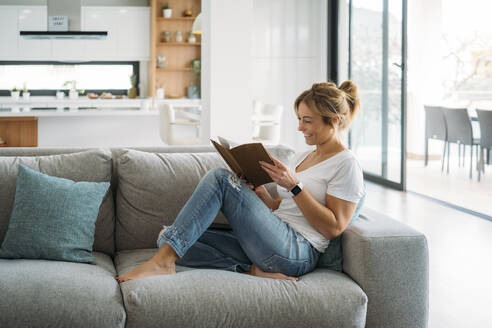 Female freelancer sitting on sofa while reading book at home - MPPF01654