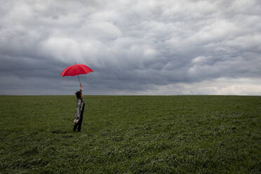 Unbekümmerte Frau mit rotem Regenschirm auf einem landwirtschaftlichen Feld bei stürmischem Wetter - FLLF00589