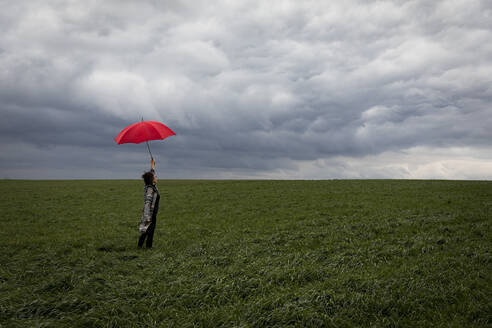 Carefree woman with red umbrella standing in agricultural field during stormy weather - FLLF00589