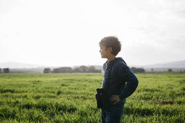 Niedlicher Junge mit Fernglas schaut weg, während er in einem Feld an einem sonnigen Tag steht - JRFF05124